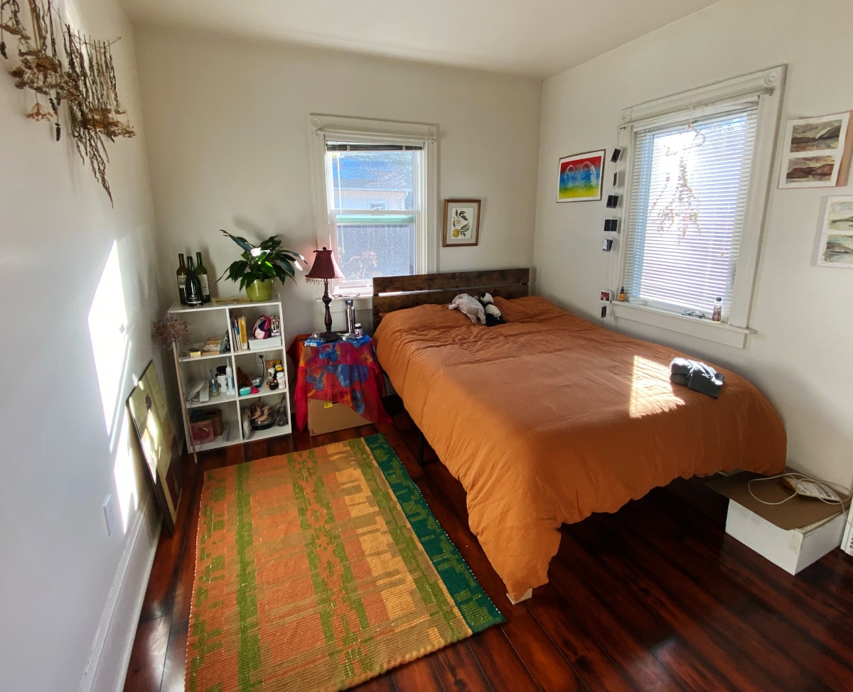 a rug on the wood floor of a sun-filled bedroom. there are prints and dried plants on the walls, and the comforter on the bed matches the rug's rust tones.