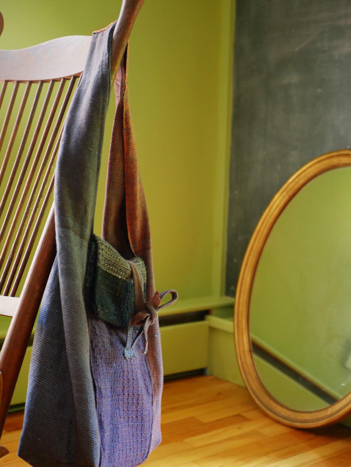 a bag hanging off the back of a rocking chair. it is boxy, with a wide shoulder strap. it has areas of purple, blue, grey, brown, and dark teal.