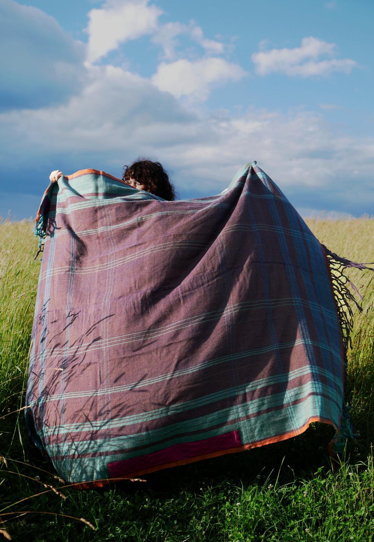 a tall photo of someone in a hay field holding up the tallit. unfolded, it billows in the wind. revealed, its stripes form a precise grid of squares within squares