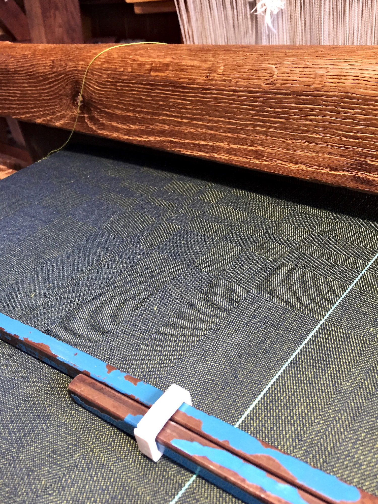 the cloth in progress on an old wooden loom. from this angle, it has blue squares slightly contrasting with more green squares.