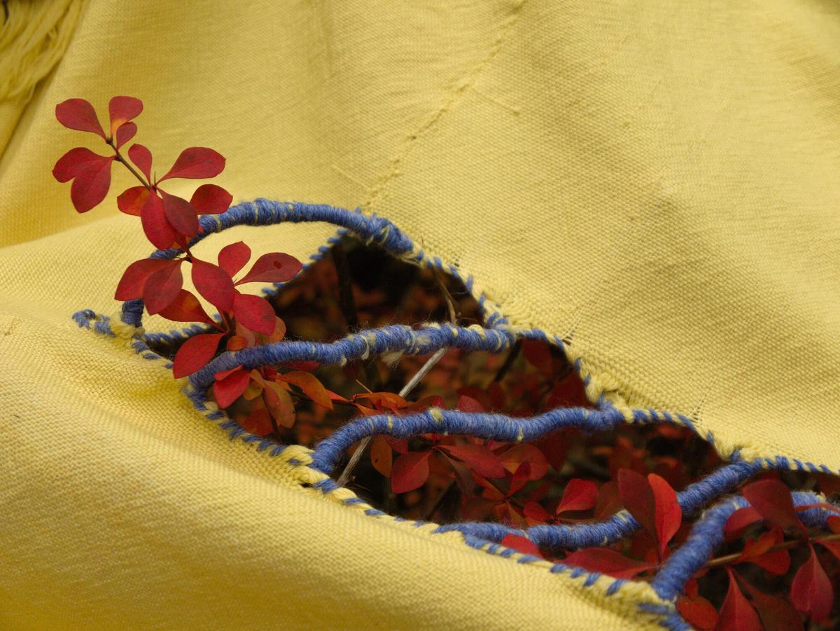 a section of cloth split into a row of open rectangles. it is held together by bridges of tightly-wrapped yarn. a red leaf pokes through.