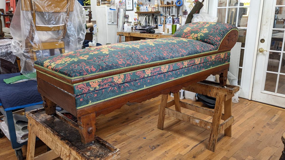 daybed in an upholstery workshop, perched on sawhorses. it has a dark wood frame, elaborate green and red floral velvet covering, and each cushion is lined with green and yellow trim.